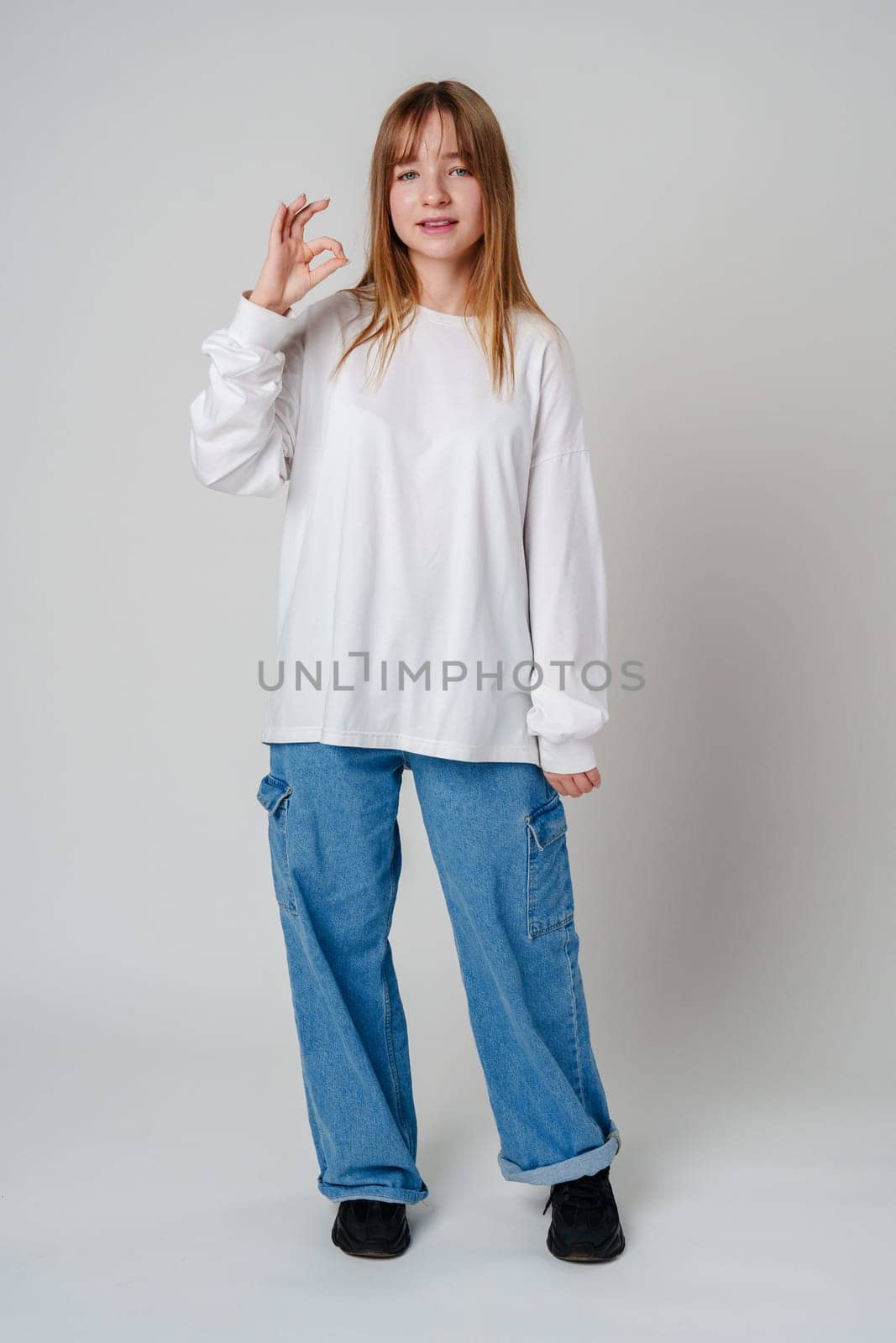 Casual young woman posing confidently in a white shirt and blue pants against a neutral background indoors