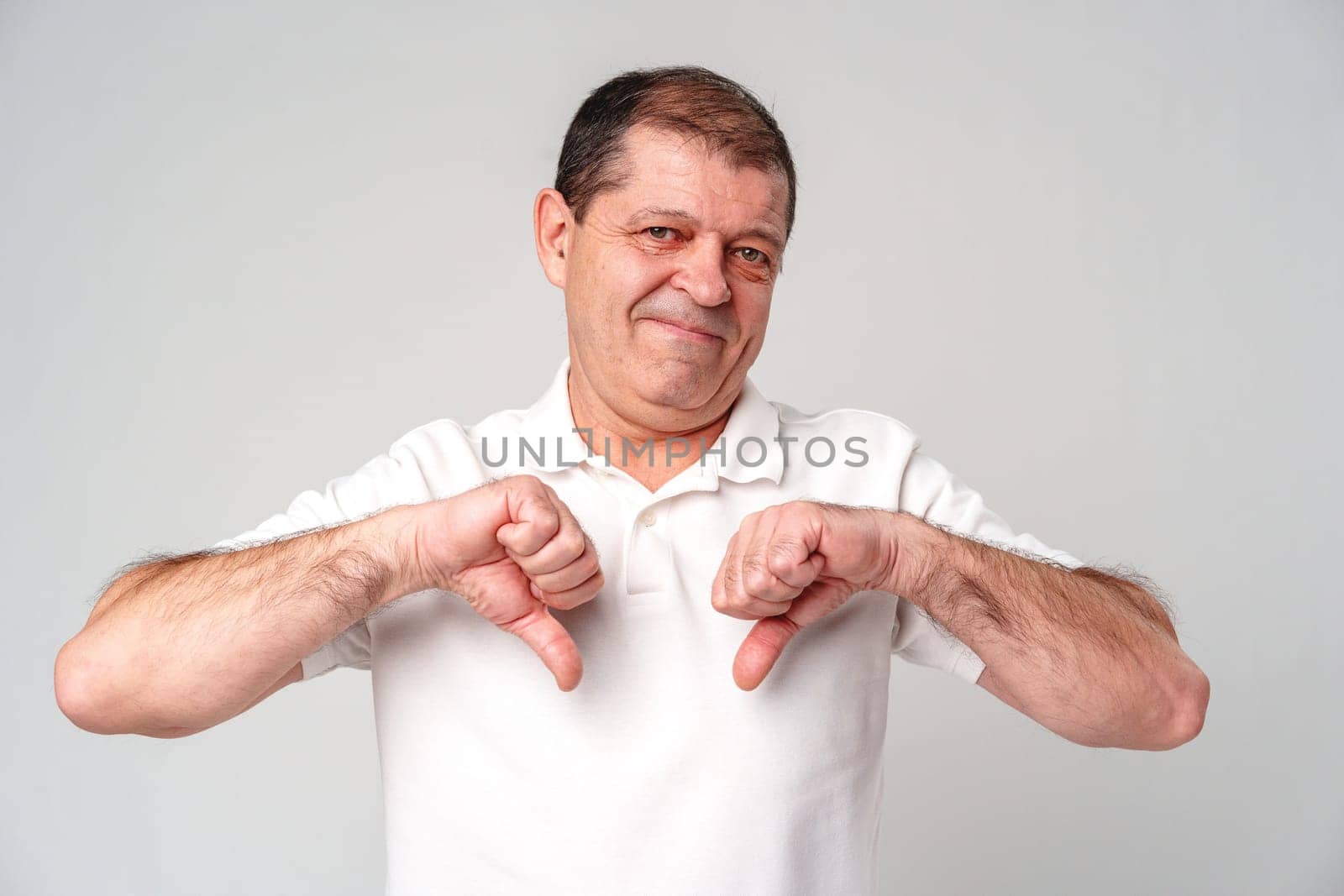 Older man expressing disapproval with thumbs down gesture while standing against a plain background close up
