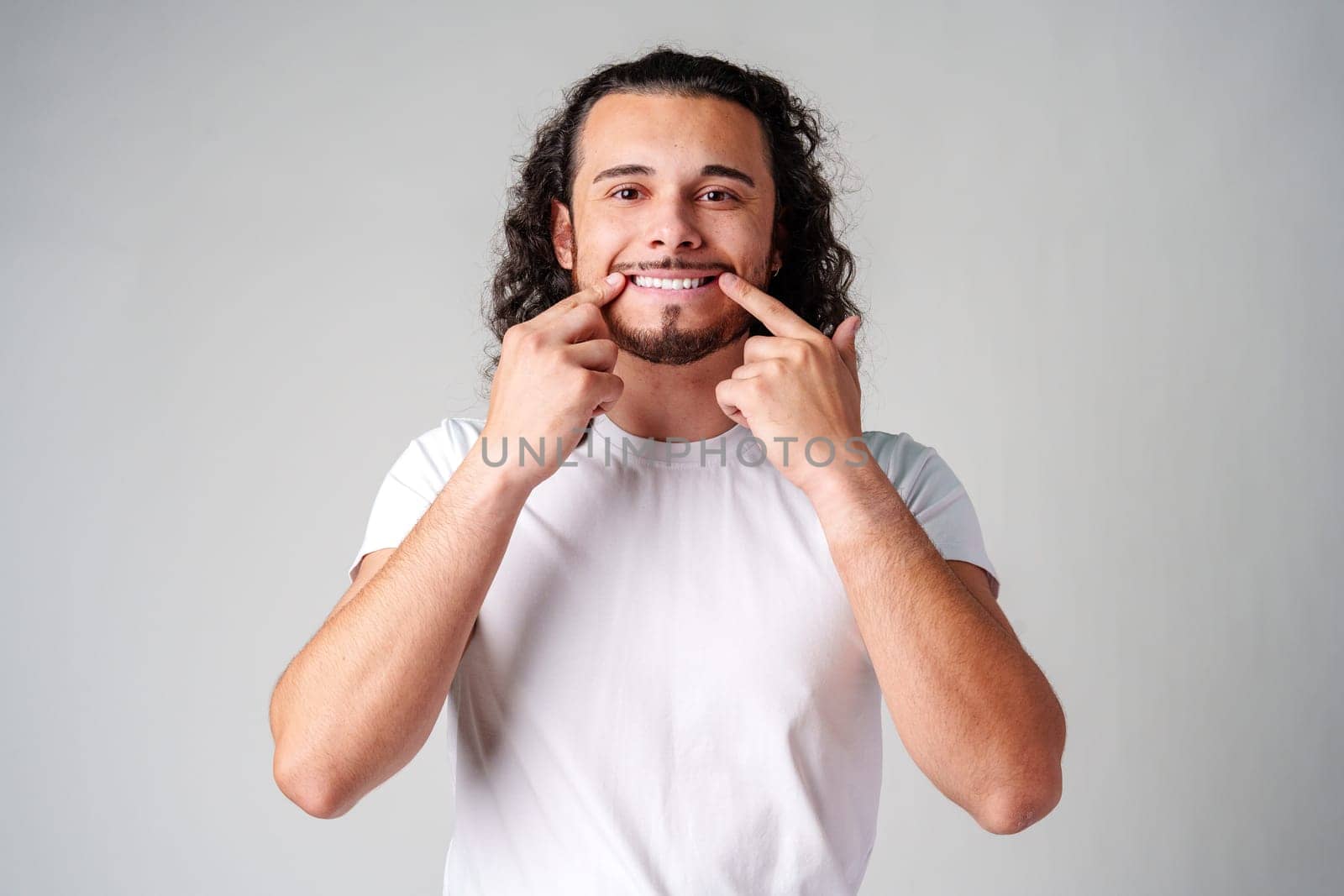 Young man in a white t-shirt playfully smiles while pointing to his teeth against a plain gray background in a studio setting