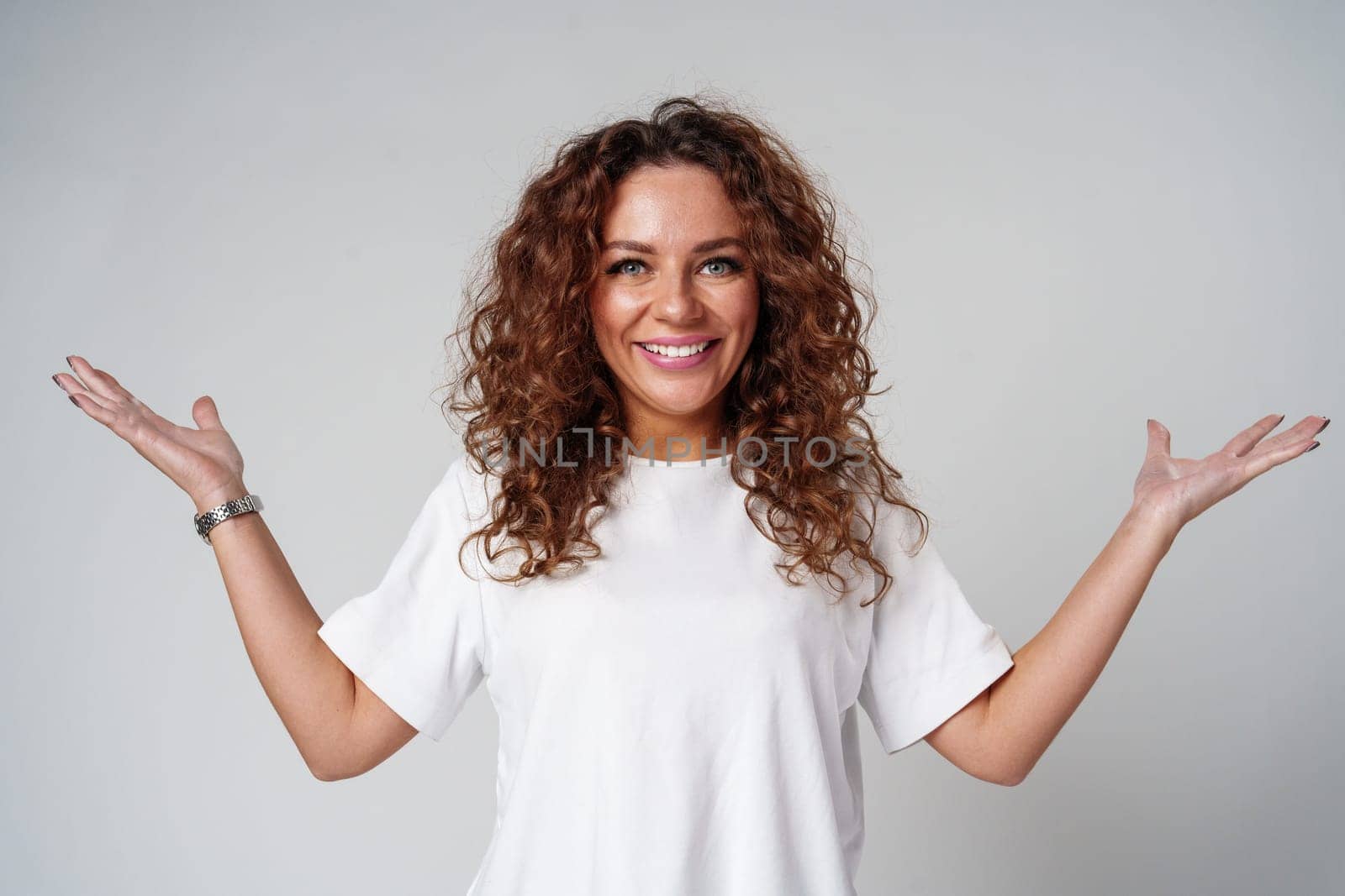 Cheerful woman with curly hair poses with open arms in a white shirt against a plain background close up
