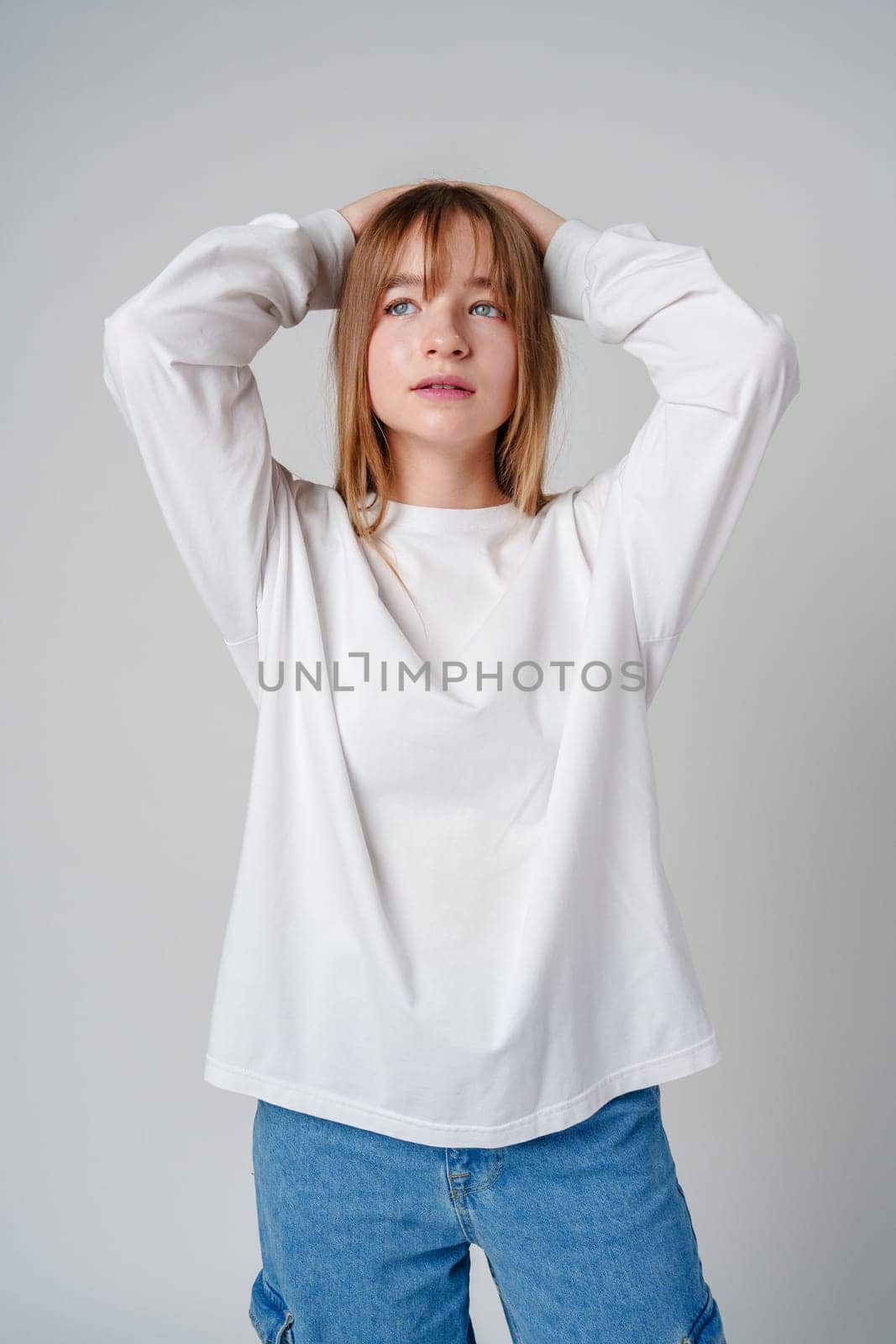 Young woman in a casual white long-sleeve shirt and loose denim pants poses against a light gray background in a studio setting