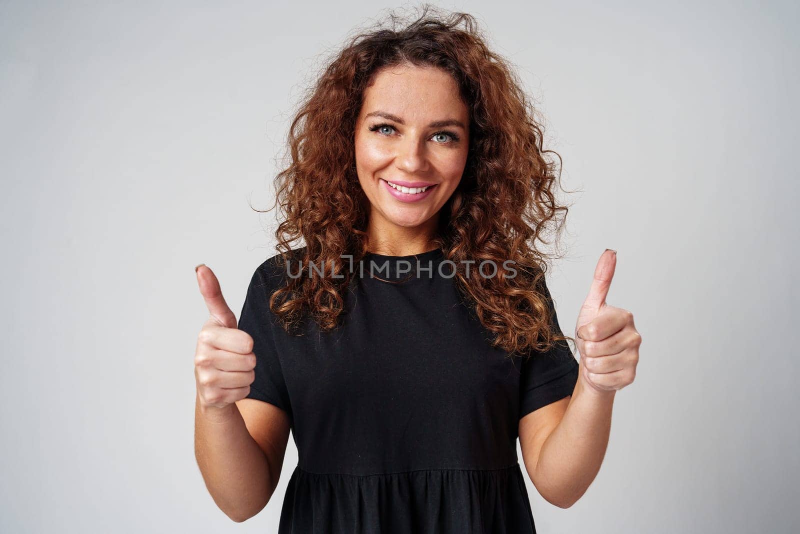 Cheerful young woman with curly hair giving a thumbs up while smiling against a neutral background close up