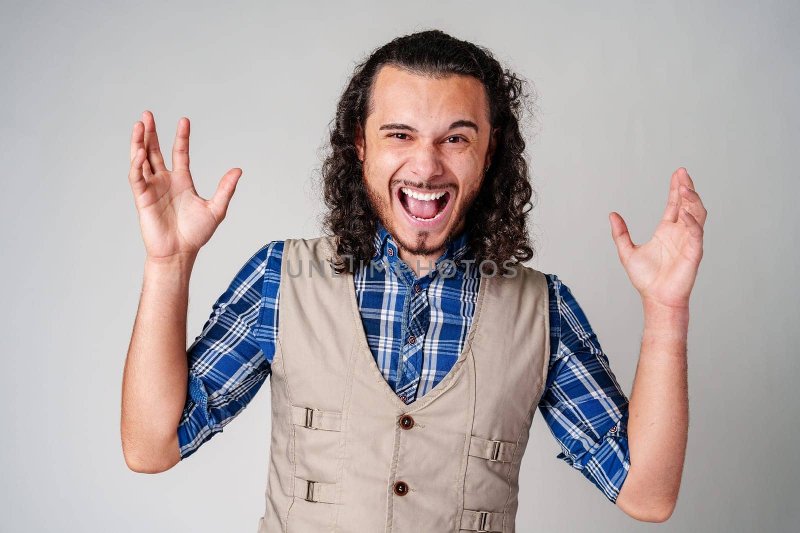A man with curly hair excitedly gesturing while wearing a plaid shirt and vest against a plain background close up