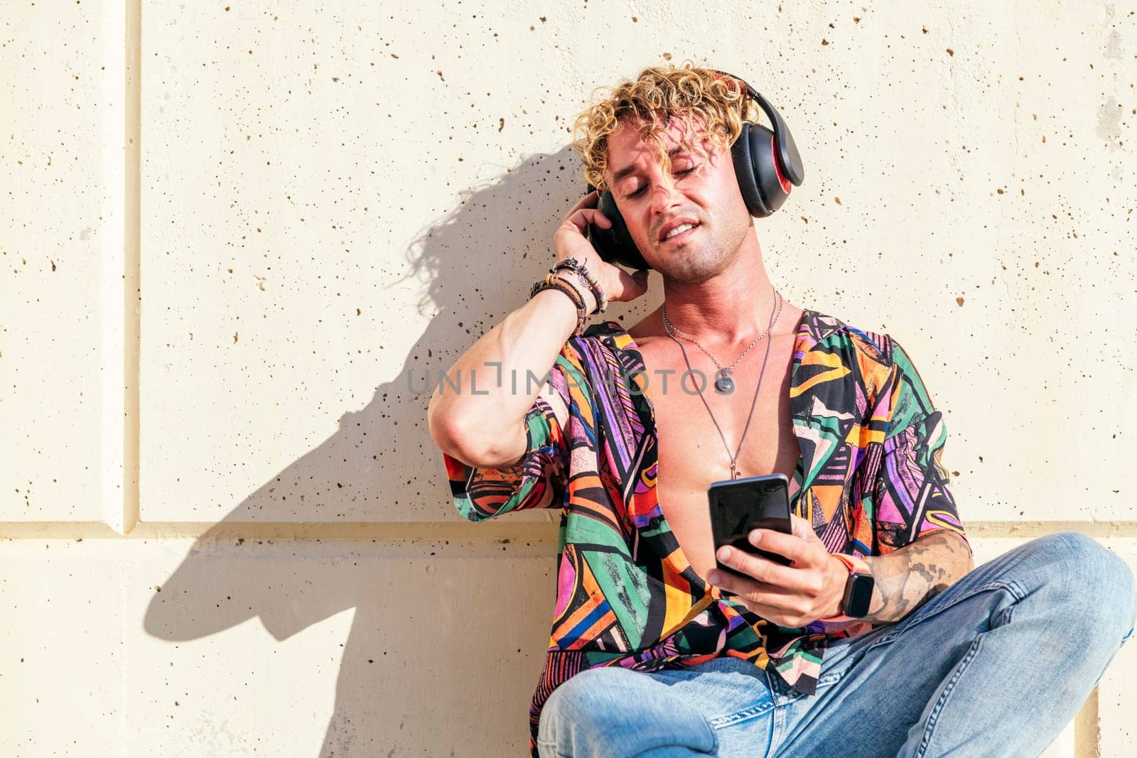 male hipster sitting on street and enjoying songs in headphones with closed eyes