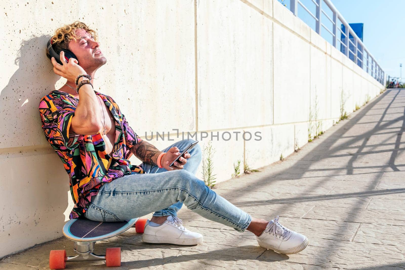 side view of dreamy male hipster sitting on skateboard on street and enjoying songs in headphones with closed eyes