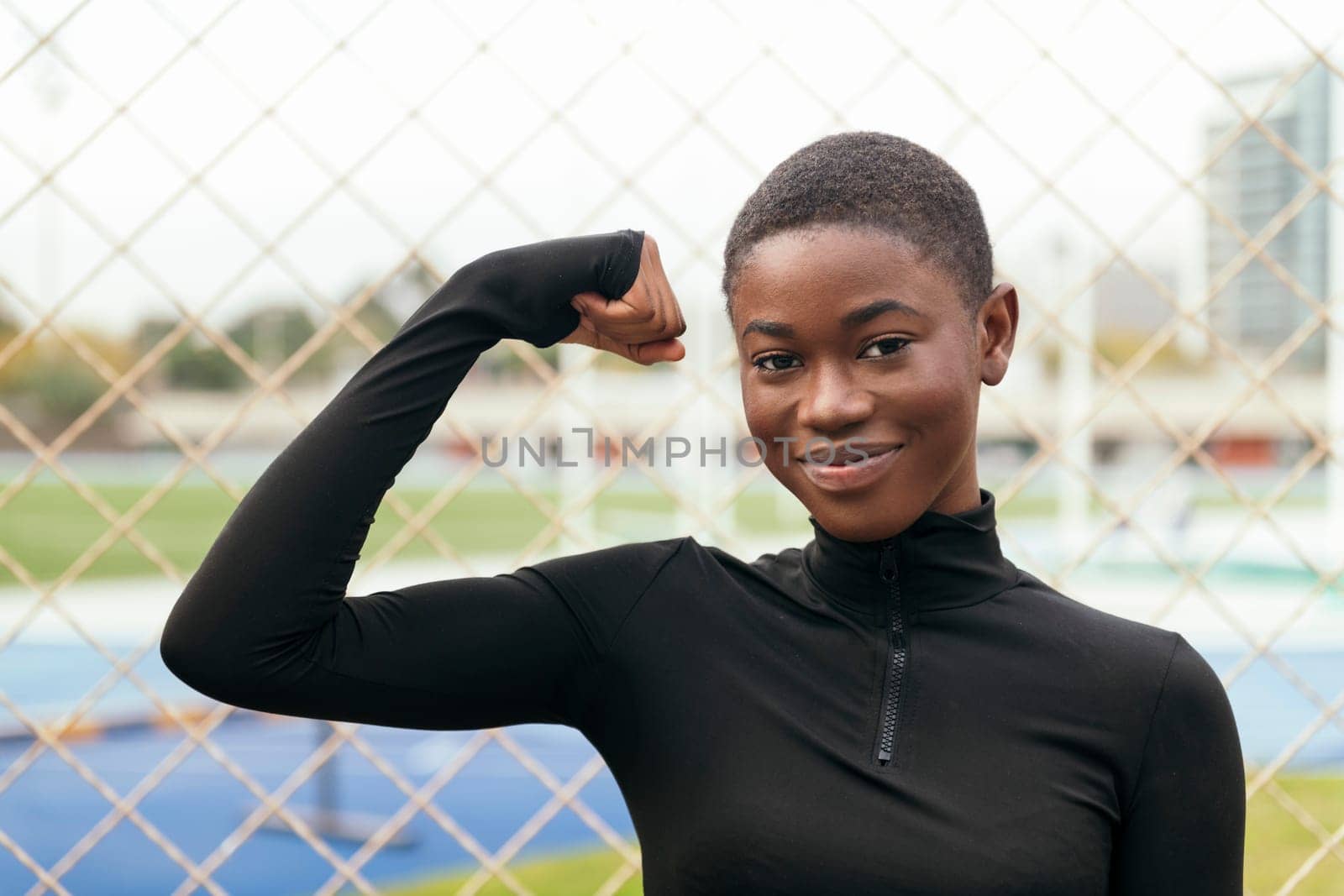 young content african american female with short hair looking at camera while raising arm showing muscle against grid fence
