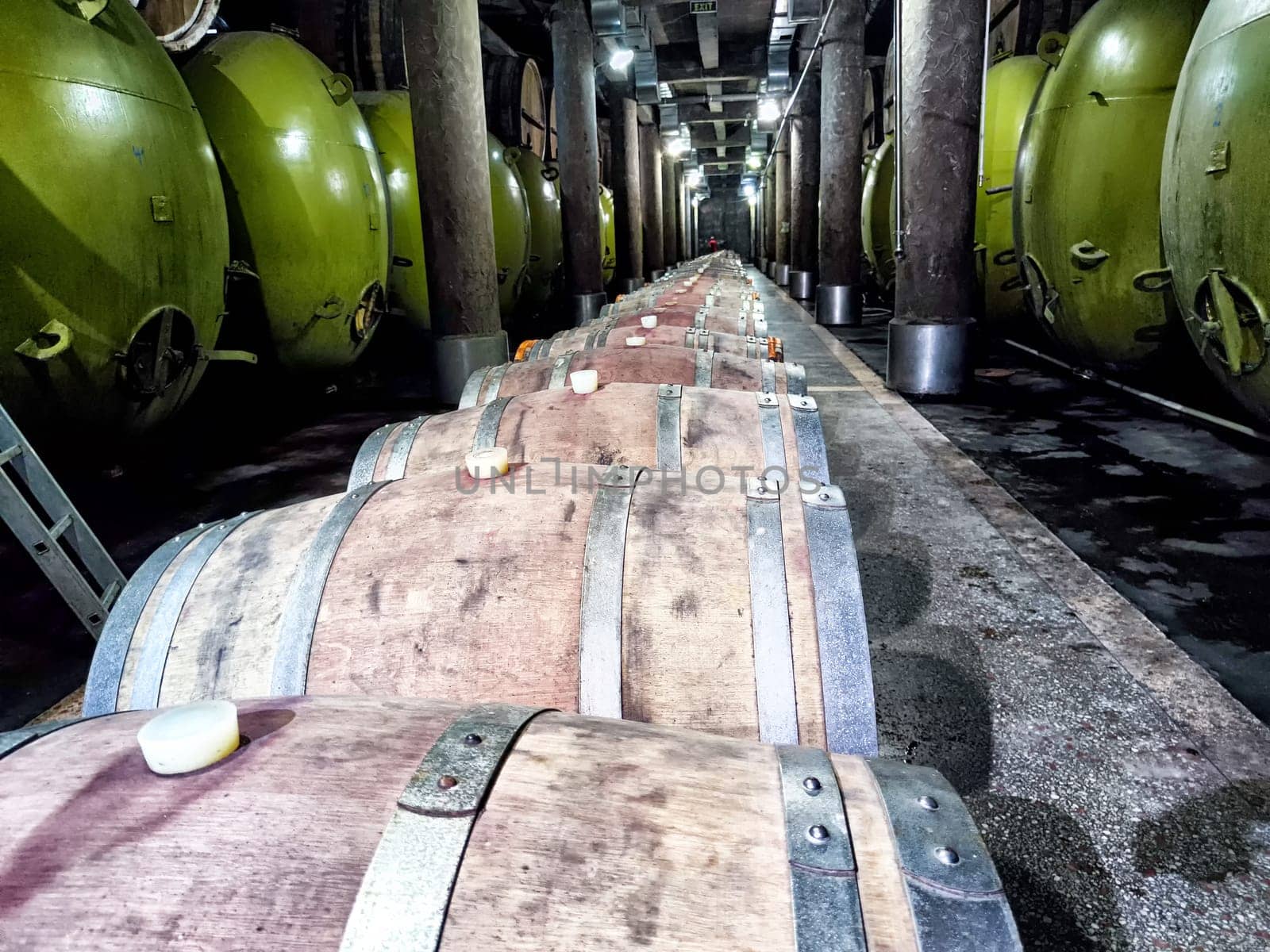 Inside the wine cellar at Kindzmarauli Winery, showcasing a row of oak barrels and green fermentation tanks in a dimly lit environment.