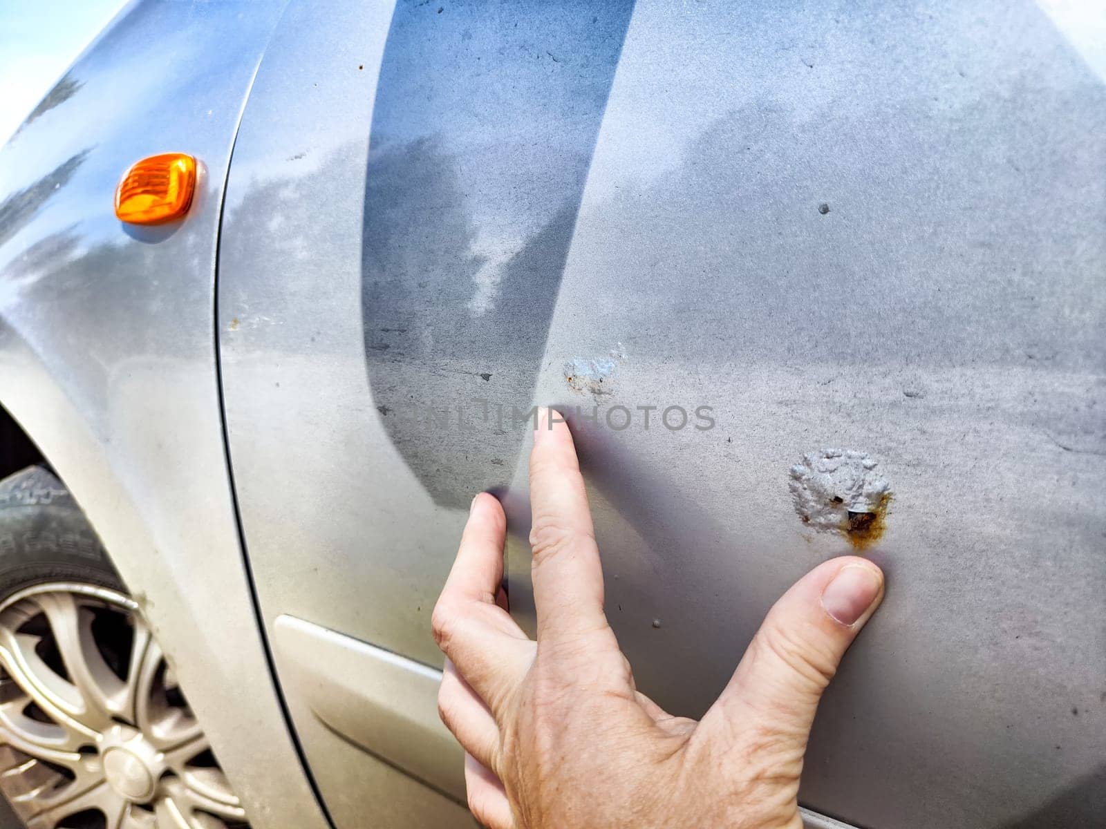 A person examines rust and scratches on silver car parked in a bright outdoor setting, focusing on the damaged area