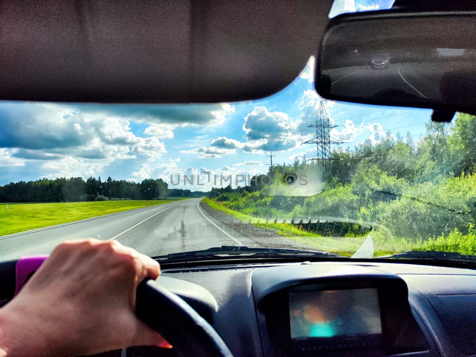 The driver steers along highway, with vibrant green fields and dramatic clouds stretching across the sky