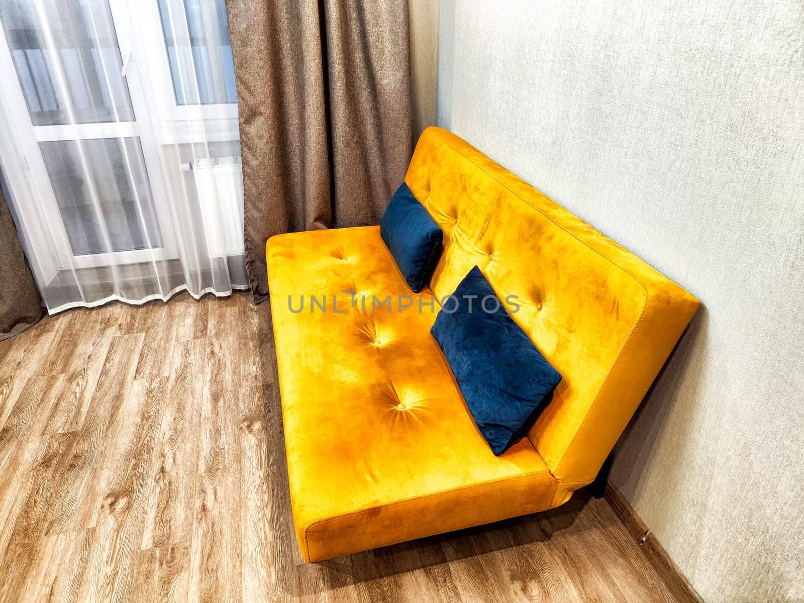 A vibrant orange sofa with blue cushions sits against a light wall in a sunny living space.
