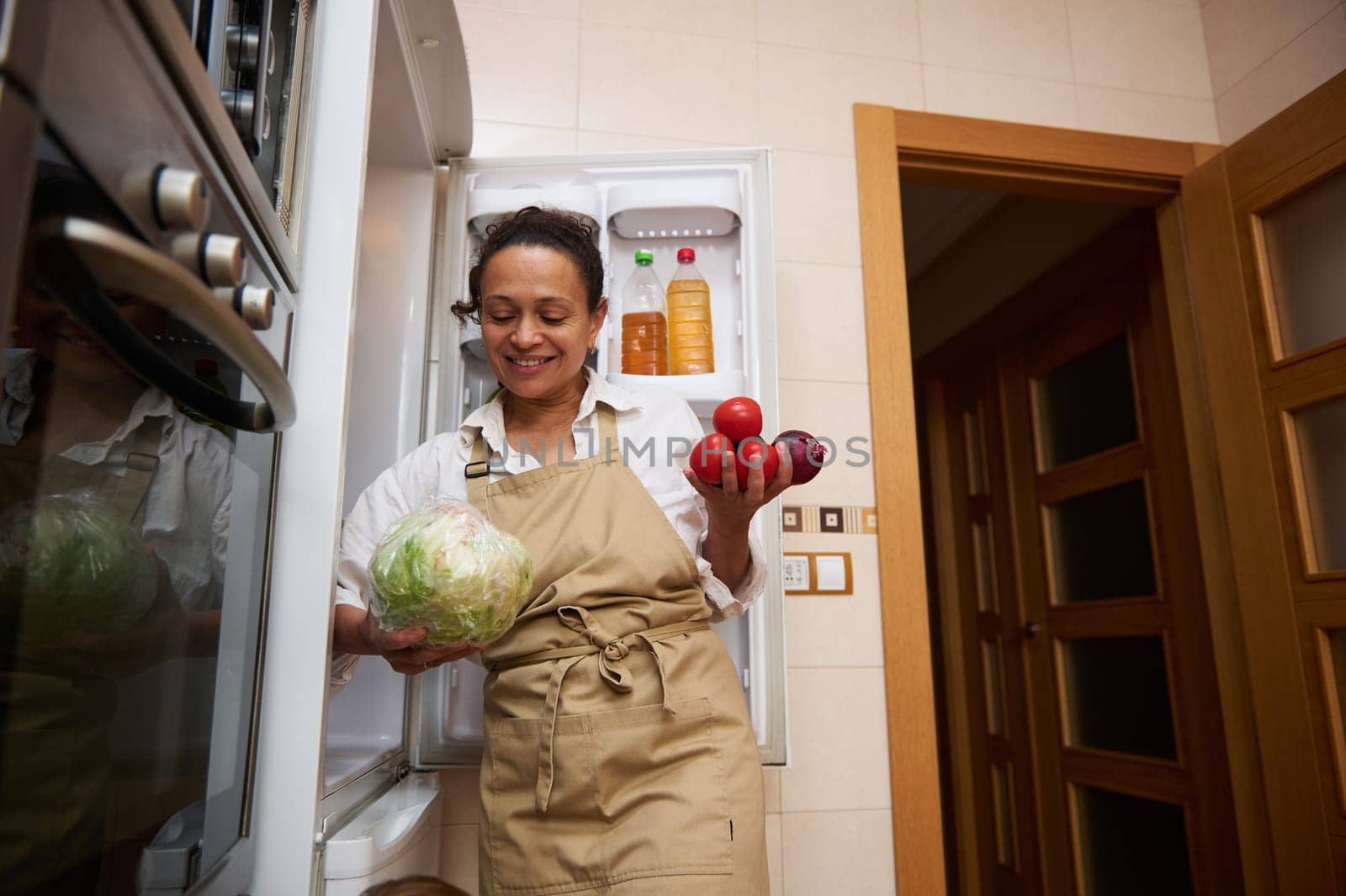 A smiling woman in an apron stands in her kitchen, holding lettuce and tomatoes, while organizing her fridge. The scene conveys a sense of healthy living and home cooking.