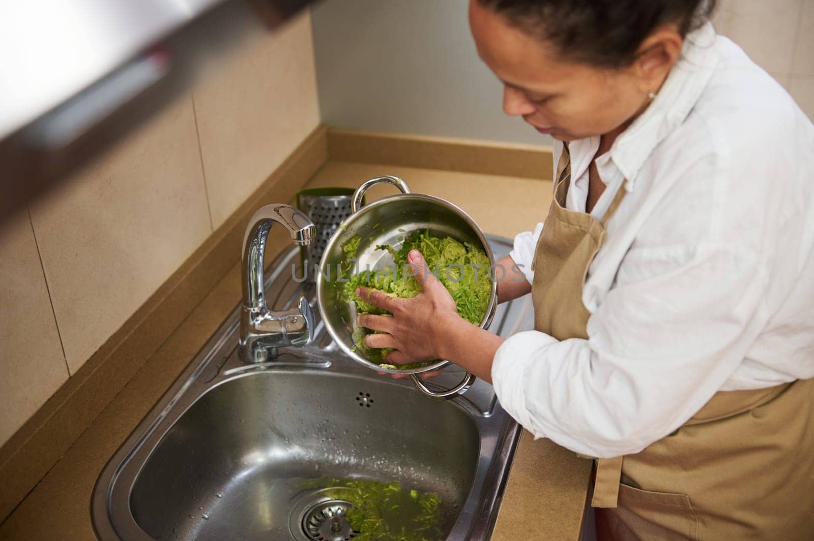 A woman in an apron washes fresh green vegetables in a kitchen sink. The scene conveys cleanliness and preparation, focusing on healthy eating and daily chores in a cozy kitchen setting.