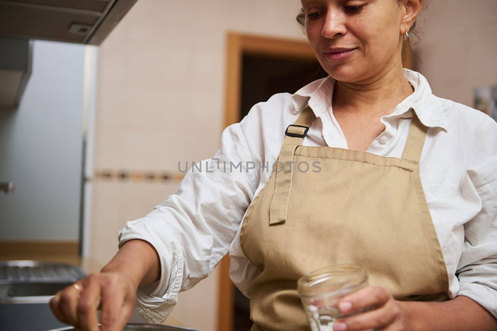 A woman in a beige apron is cooking in a home kitchen, carefully preparing a dish with a mason jar in hand, reflecting domestic warmth.
