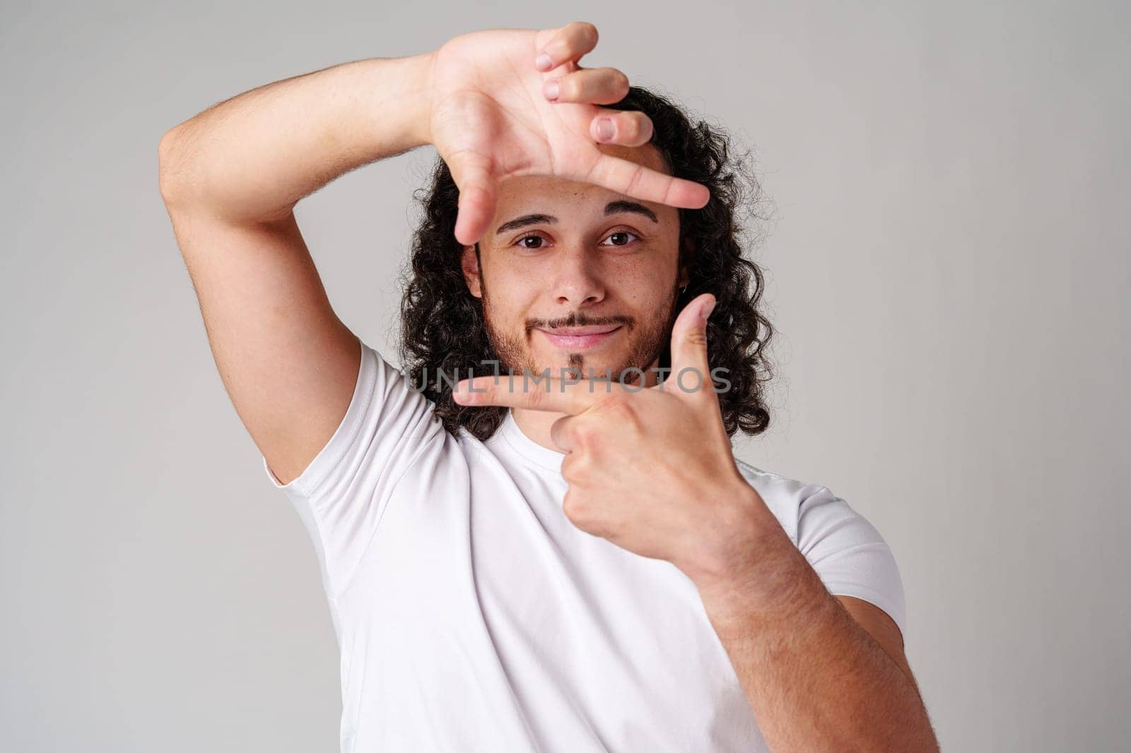 A young man with curly hair framing his face and making a gesture as if capturing a moment against a plain background close up