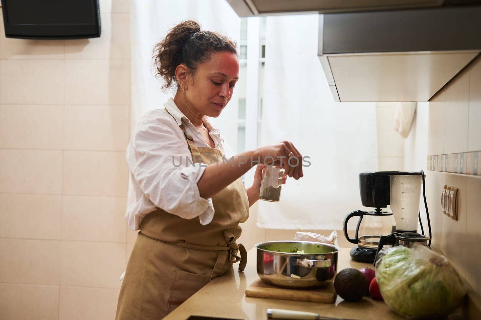 A woman in an apron adds seasoning to a fresh salad in a modern kitchen. The setting is cozy and bright, with a focus on healthy cooking and home lifestyle.