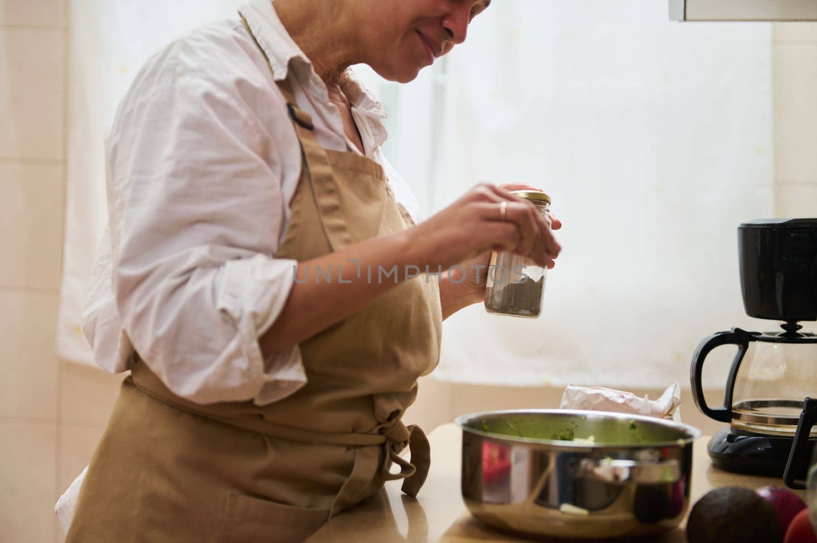 Woman in apron seasoning food in a home kitchen, creating a homemade meal with fresh ingredients, showcasing culinary skills and a love for cooking.