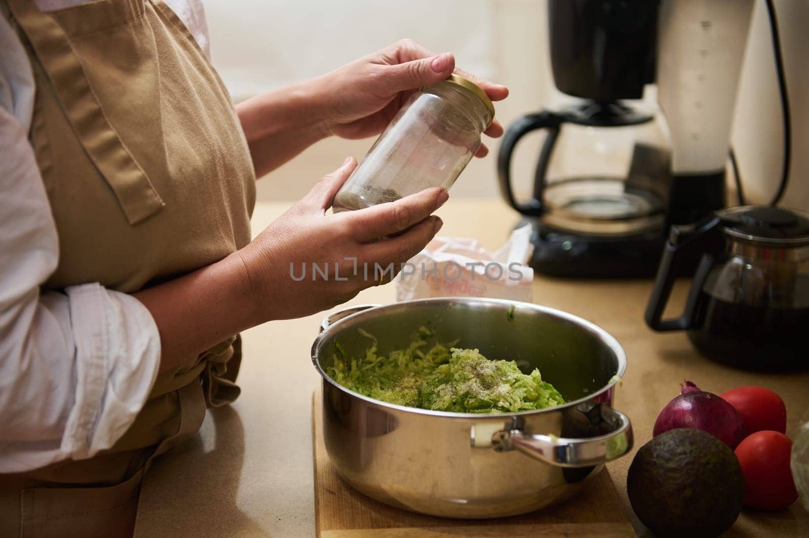 A person in an apron adds spices to a pot of fresh greens on a kitchen counter. Nearby are tomatoes, an onion, and an avocado, indicating a focus on homemade and healthy cooking.