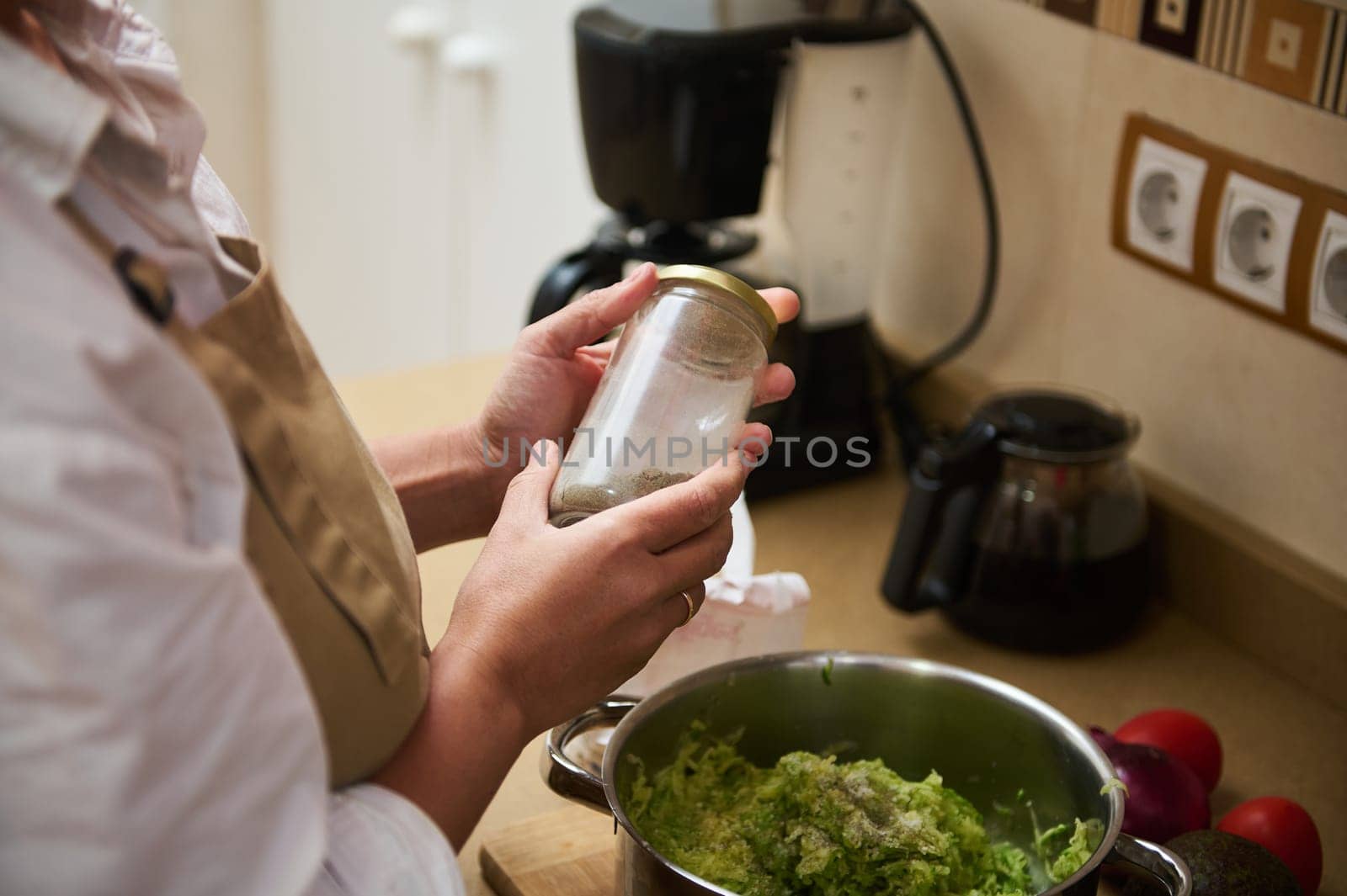 A woman in an apron seasons a dish in a home kitchen, adding a personal touch to her culinary creation. Fresh ingredients are prepared on the counter, reflecting home cooking passion.