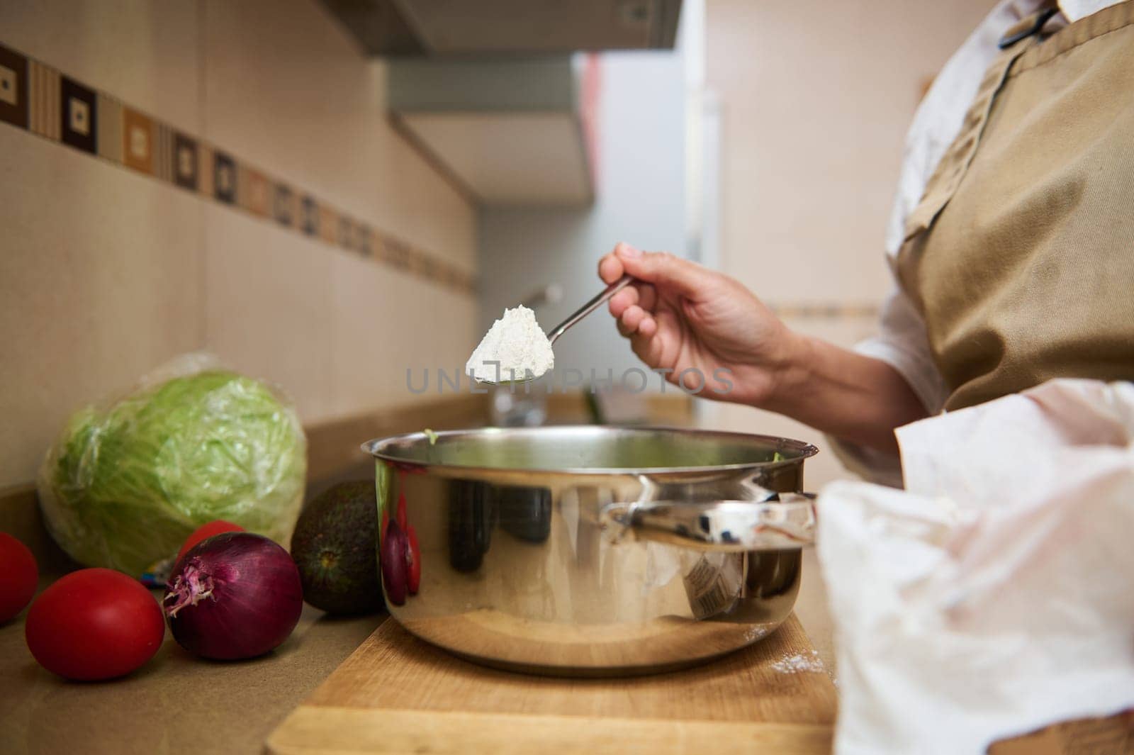 A person in an apron adds ingredients to a pot in a cozy kitchen setting, surrounded by fresh vegetables. The image captures the essence of home cooking and the joy of preparing meals.