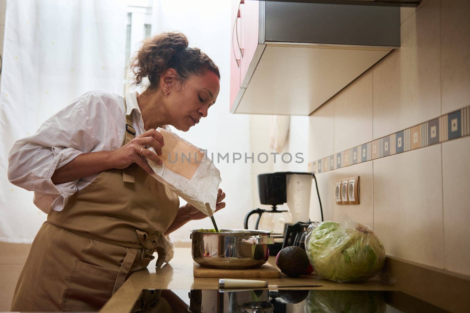 A woman in an apron is seen cooking with fresh vegetables, pouring ingredients into a pot in her modern kitchen. The scene exudes warmth and creativity in home cooking.