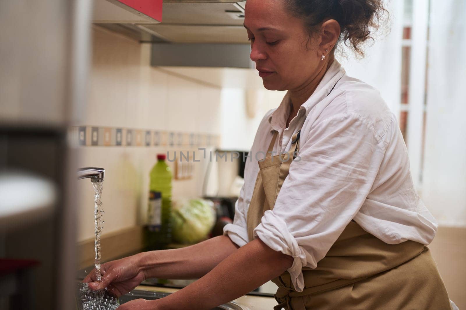 A woman wearing an apron washes her hands at the kitchen sink, ensuring cleanliness and hygiene. The running water and kitchen setting emphasize a focus on healthy home practices.