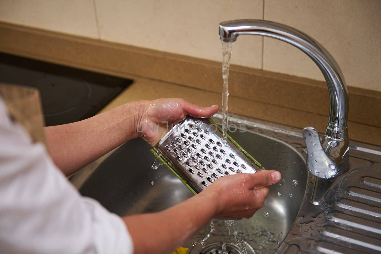 Close-up of hands cleaning a metal grater under running tap water. The process of washing kitchen utensils after use, promoting cleanliness and hygiene.