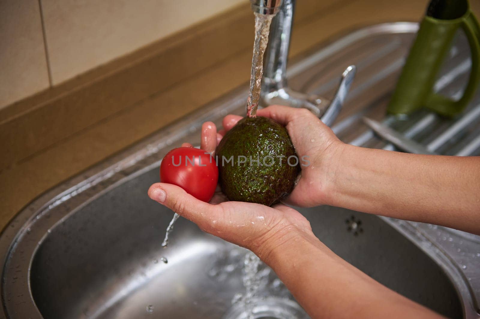 Close-up of hands washing a fresh avocado and tomato under running water in a kitchen sink, promoting cleanliness and food hygiene.