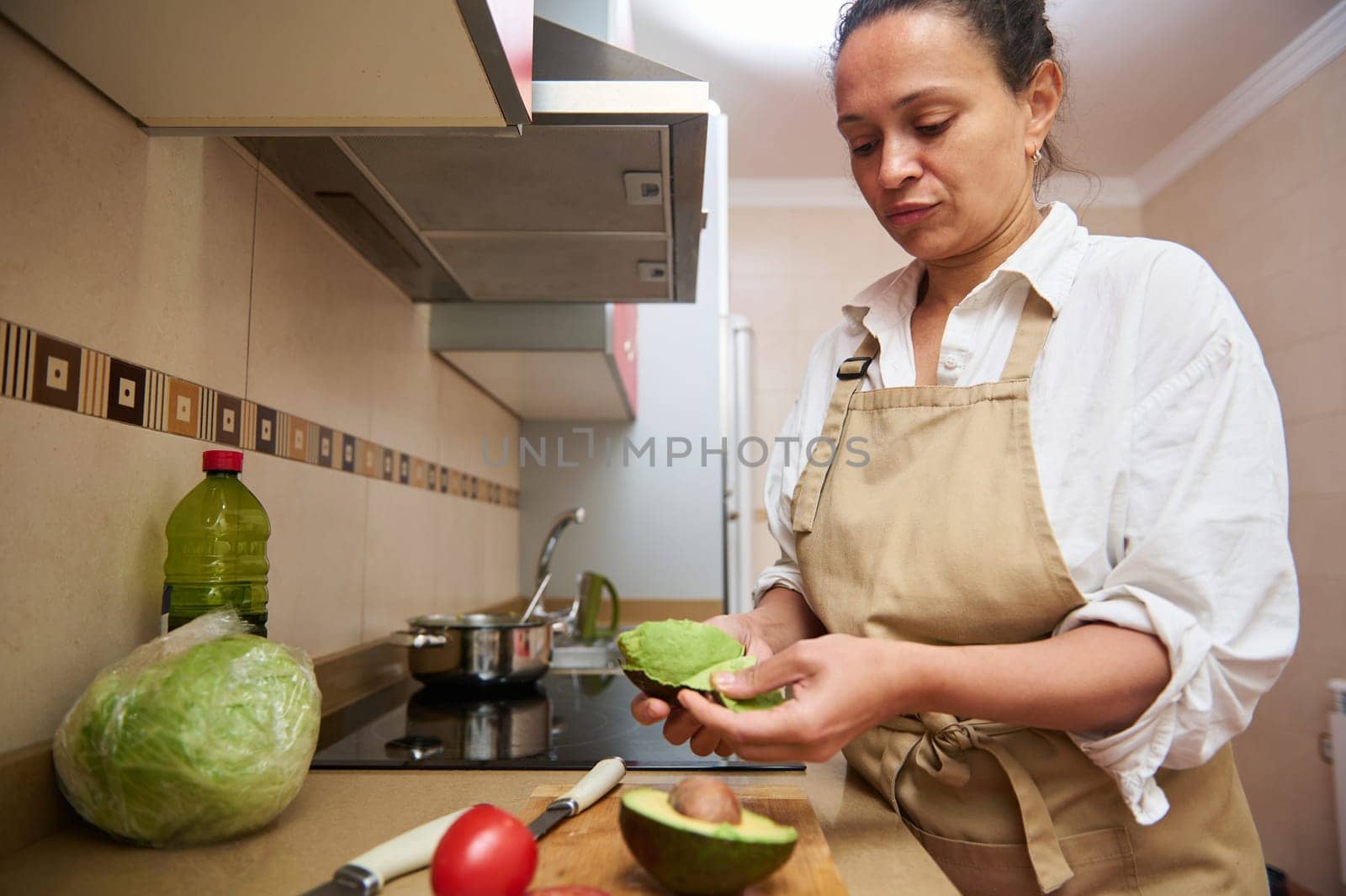 A woman in a kitchen carefully peeling an avocado, surrounded by fresh ingredients. She wears an apron, suggesting a focus on healthy cooking and meal preparation.