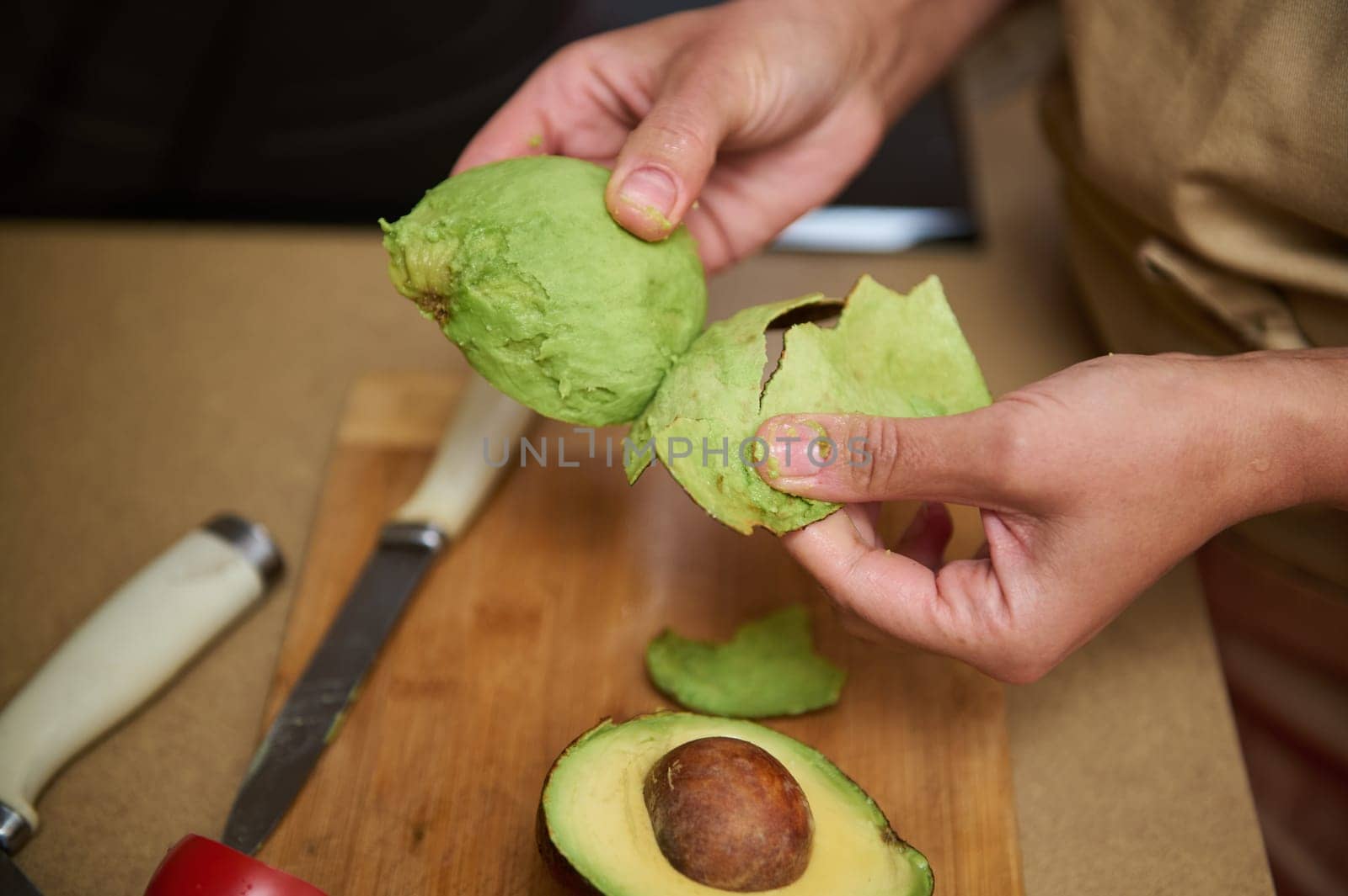 Close-up of hands peeling an avocado on a wooden cutting board. A whole avocado is sliced open, showcasing its vibrant green flesh and large seed, emphasizing freshness and culinary preparation.