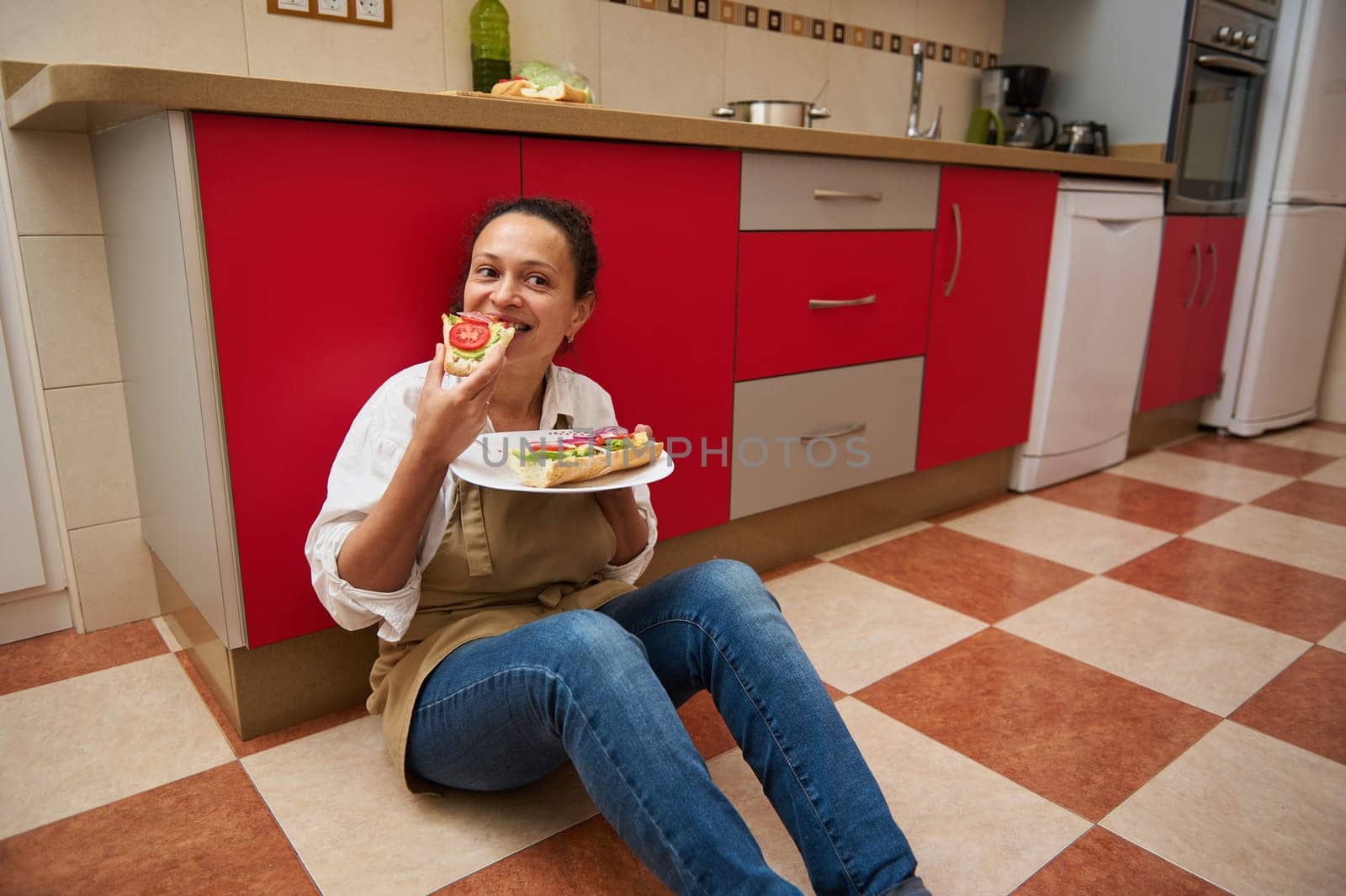A cheerful woman sits on a kitchen floor, enjoying a homemade sandwich. She is relaxed with a joyful expression, showcasing a casual and content moment in a home environment.