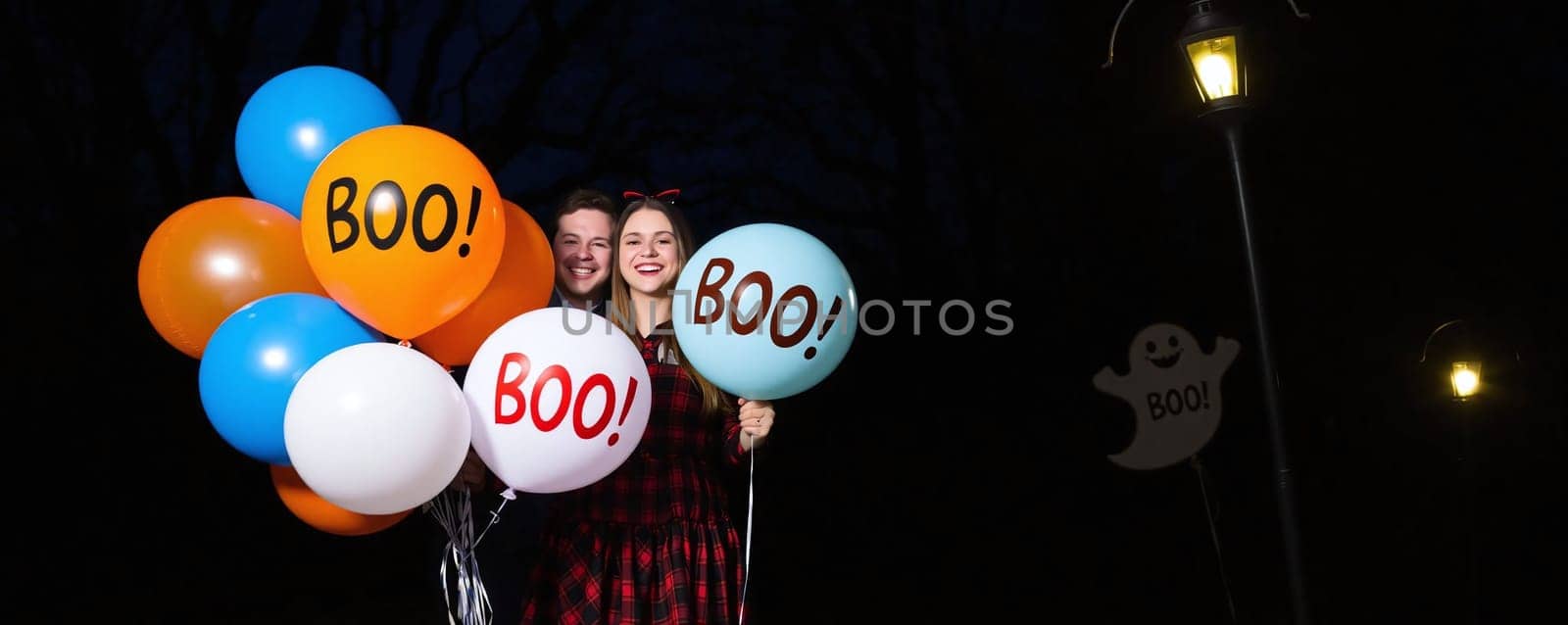 A cheerful young couple stands in a dark setting, holding colorful balloons that read BOO! The festive atmosphere captures the spirit of Halloween festivities and joy.