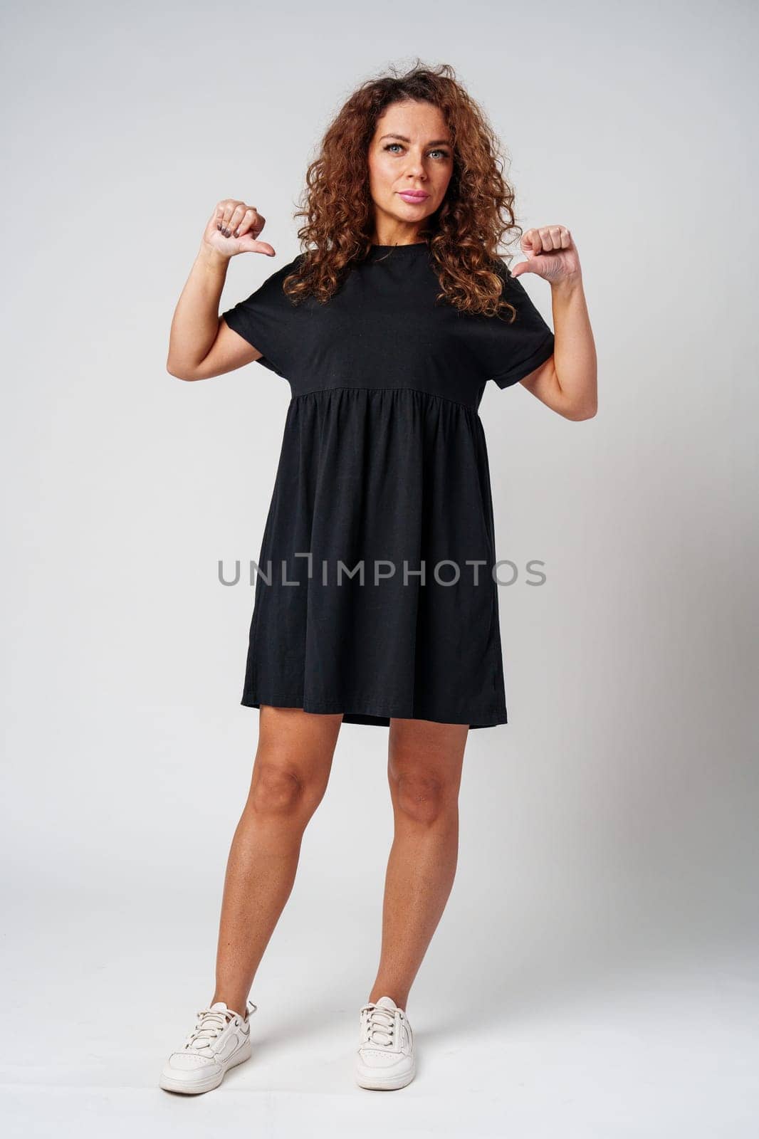 Curly-haired woman in a black dress confidently gestures downwards against a plain background close up