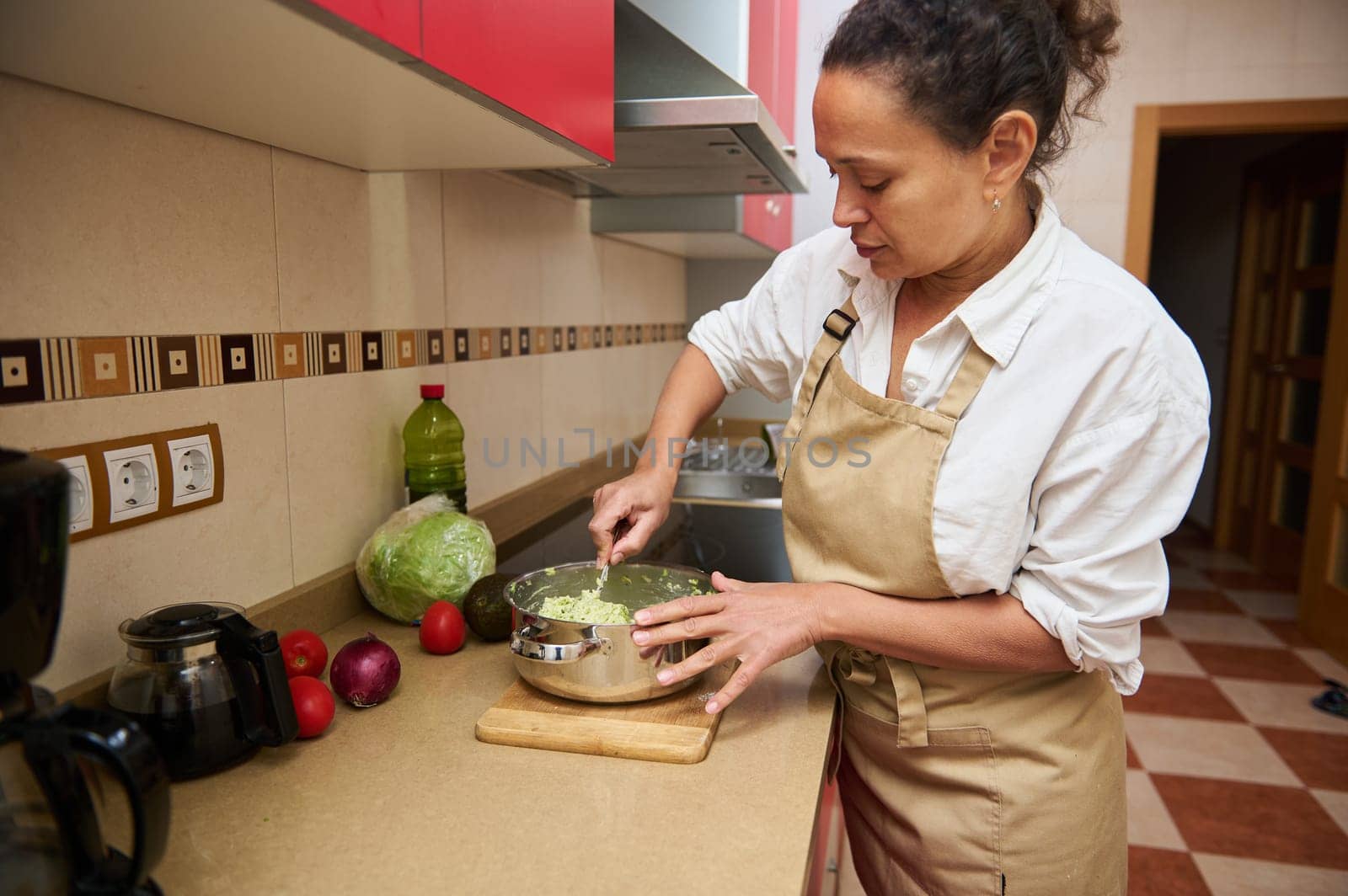 A woman in an apron mixes vegetables in a pot, preparing a fresh salad in a modern kitchen setting.