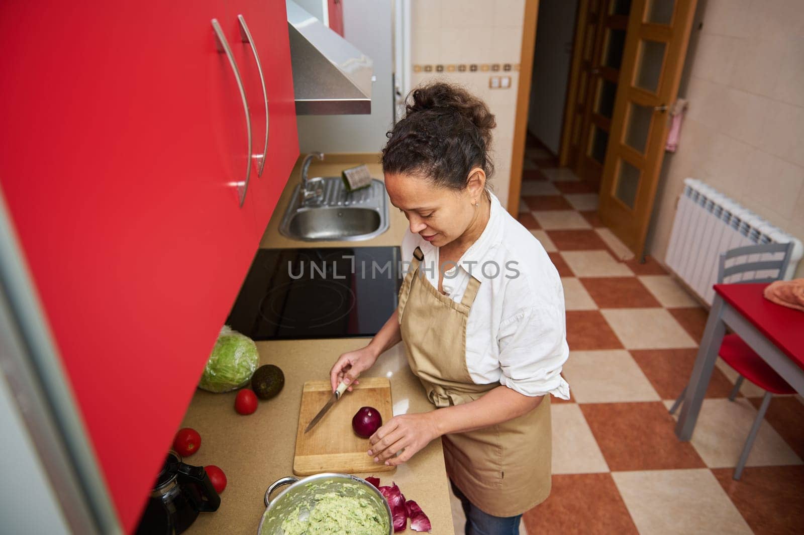 A woman wearing an apron slices an onion in a modern kitchen. Surrounded by fresh vegetables like avocados and tomatoes, she prepares a salad with focus and creativity.
