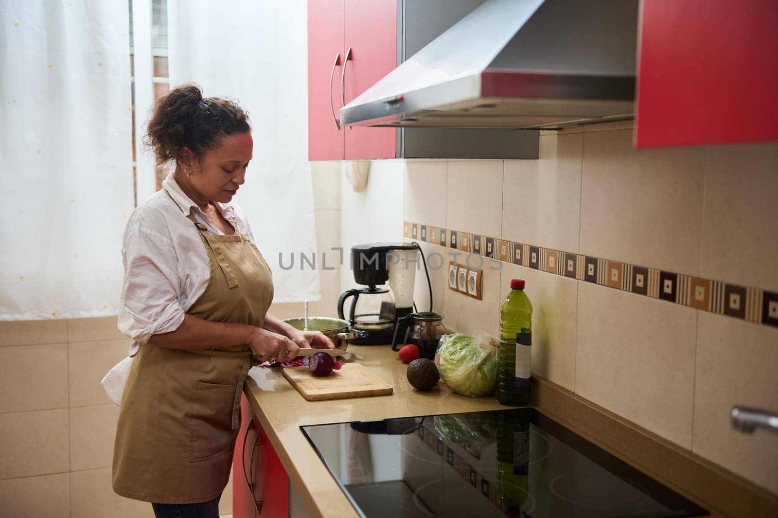 A woman wearing an apron chops vegetables on a cutting board, surrounded by fresh ingredients. The brightly lit kitchen evokes a sense of homely comfort and healthy living.
