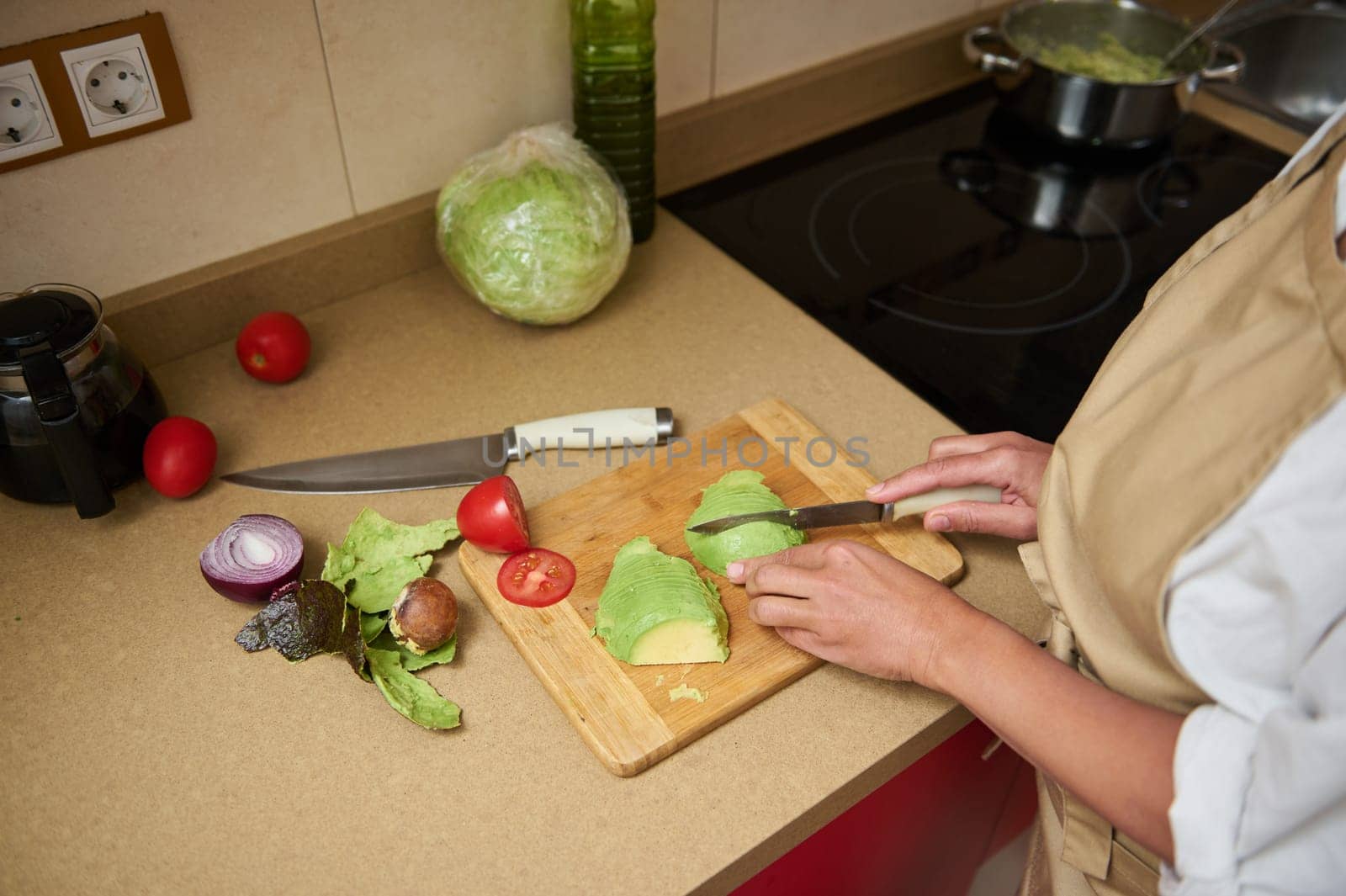 A woman slices avocado on a wooden board in a kitchen with fresh ingredients like lettuce, tomato, and onion, emphasizing healthy meal preparation.