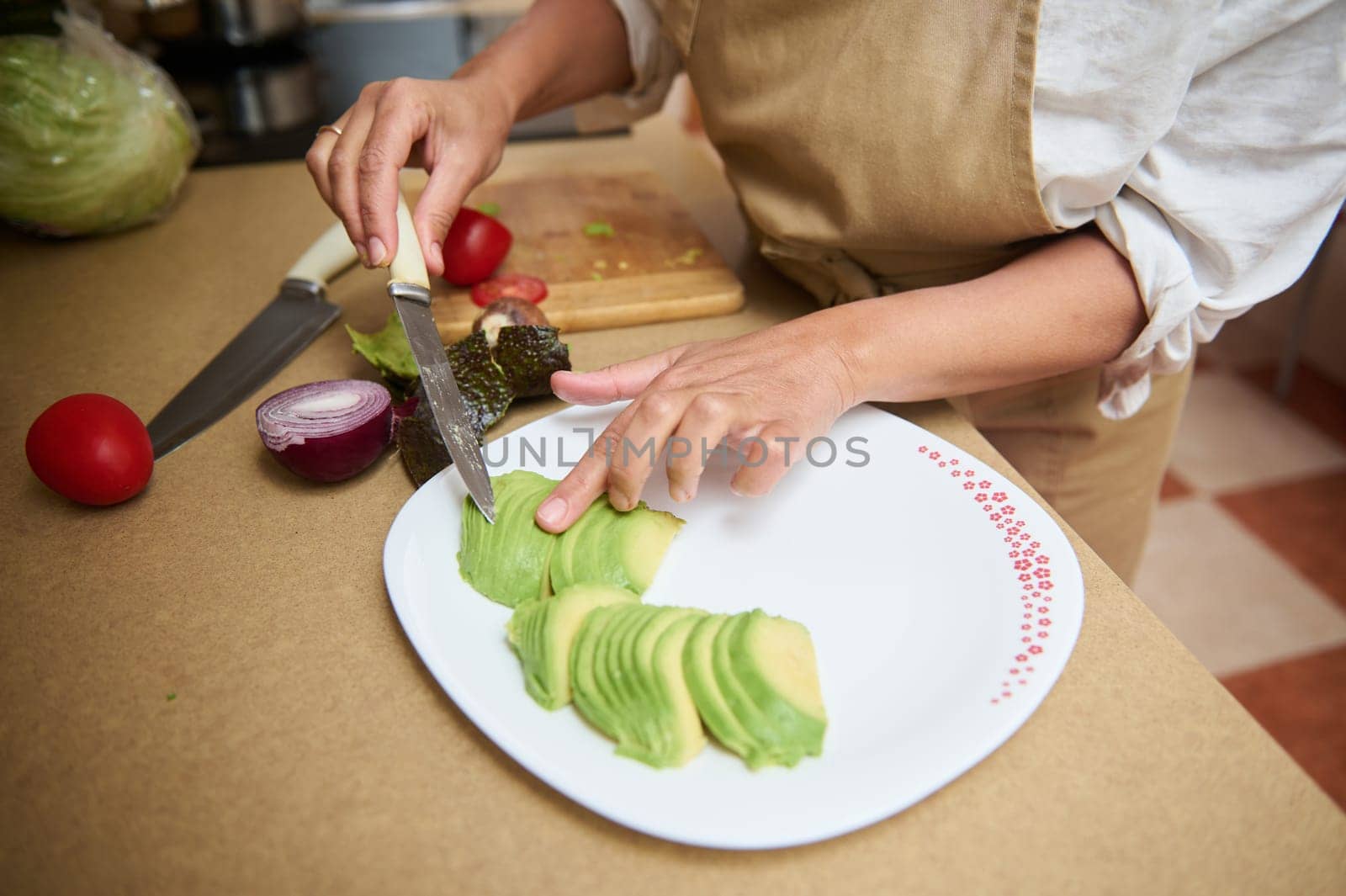 A woman is slicing ripe avocado onto a plate in a cozy home kitchen. Surrounding ingredients suggest a healthy meal preparation, with onions and tomatoes on the countertop.