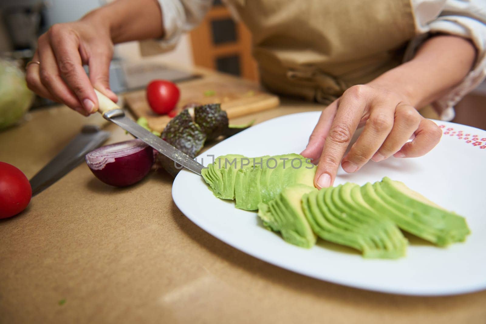 A person skillfully slices an avocado on a plate in a cozy kitchen. Fresh vegetables, including tomatoes and onions, are visible, suggesting a healthy meal preparation in progress.