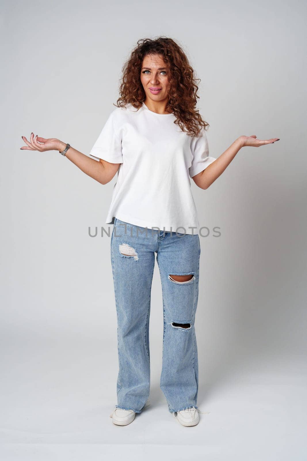Cheerful woman with curly hair poses with open arms in a white shirt against a plain background close up