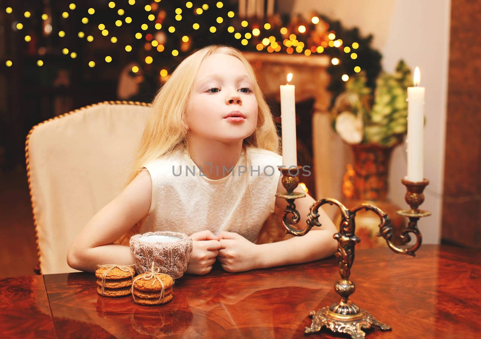 Christmas child blowing on candle makes a wish sitting at the table home