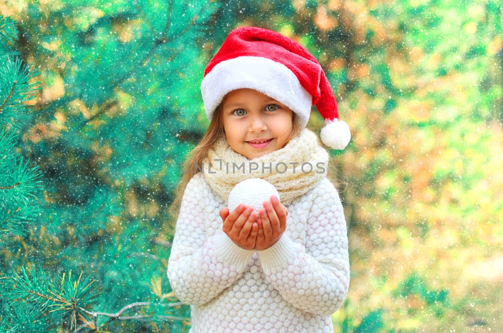 Christmas portrait smiling child little girl in santa red hat holding snowball near tree