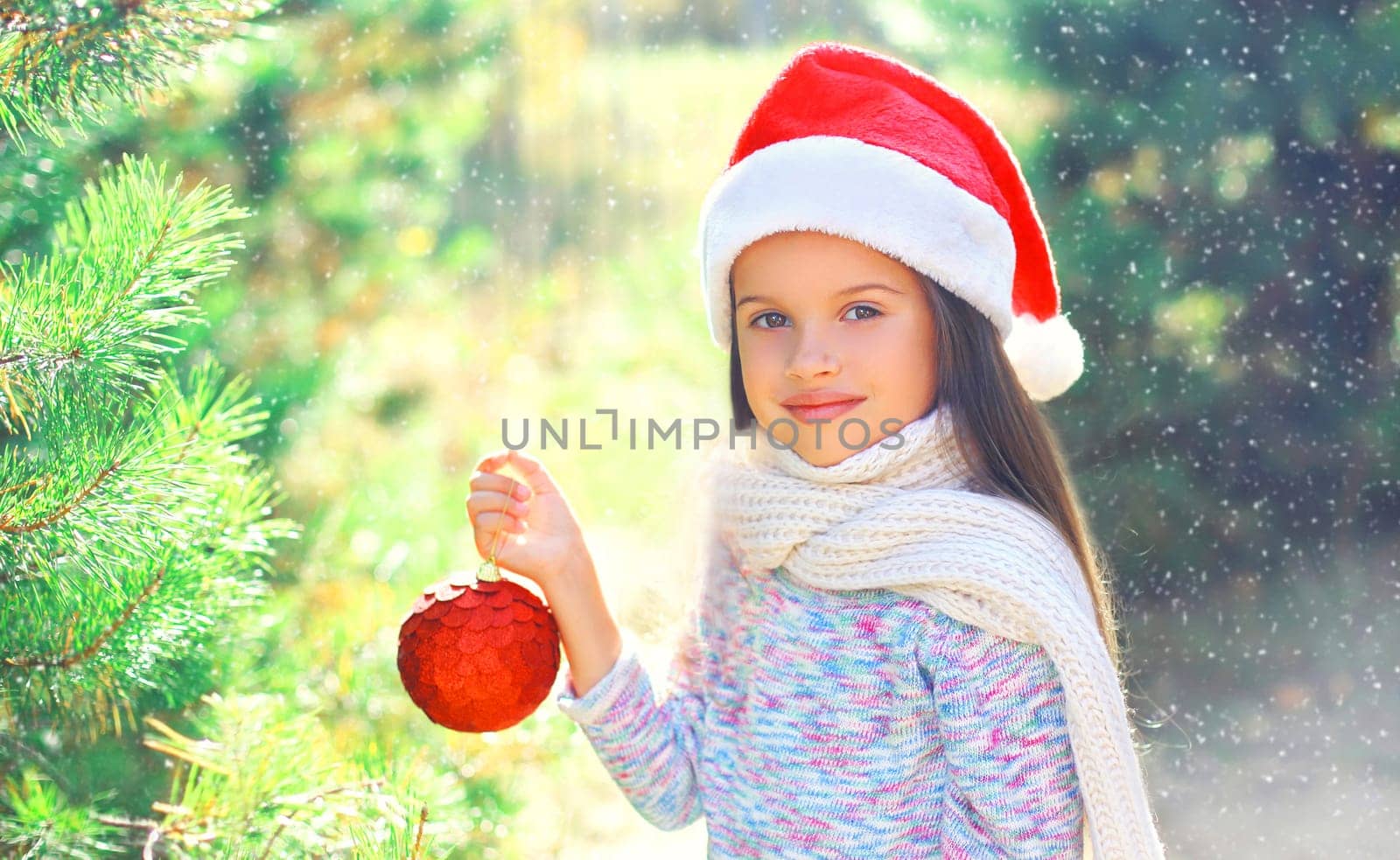 Christmas smiling child little girl in santa red hat with ball near a branch tree