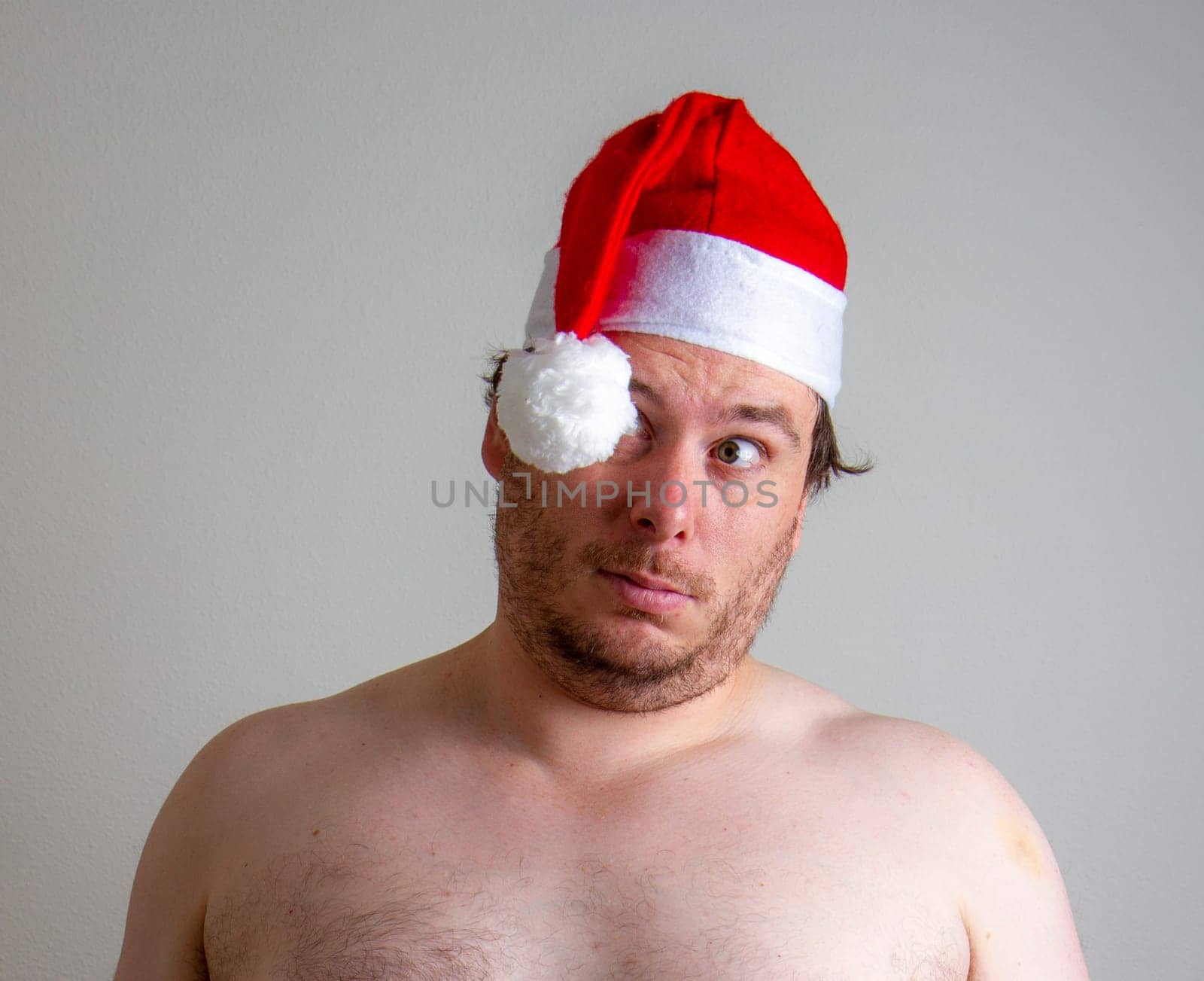 A shirtless man wearing a red Santa hat looks confused against a plain backdrop, capturing a humorous and casual holiday vibe.