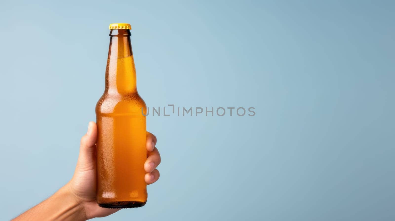A hand holds a glass beer bottle with a yellow cap against a blue background. The transparent bottle reveals the beer inside, showcasing this popular alcoholic beverage made from barley.