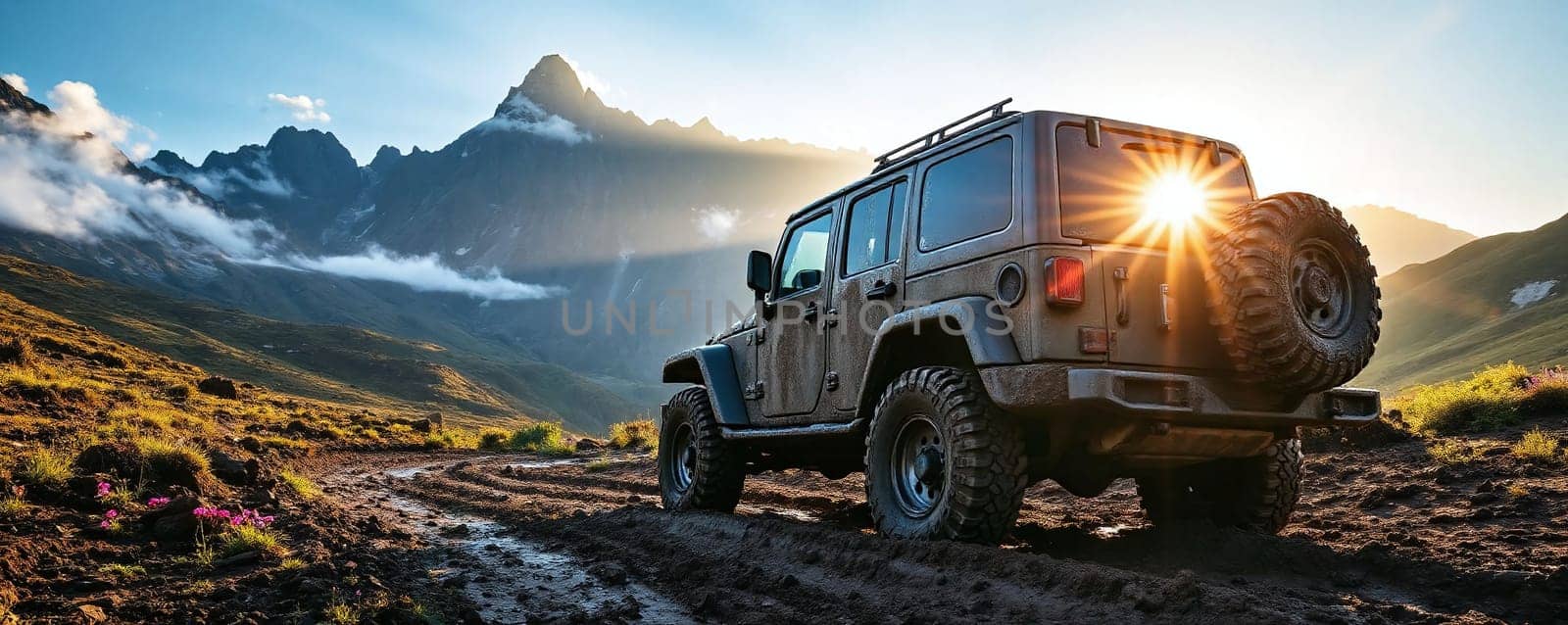 A robust off-road vehicle maneuvers along a muddy mountain trail with rugged terrain. The sun sets behind the peaks, illuminating the sky with warm hues and casting a dramatic light on the landscape.
