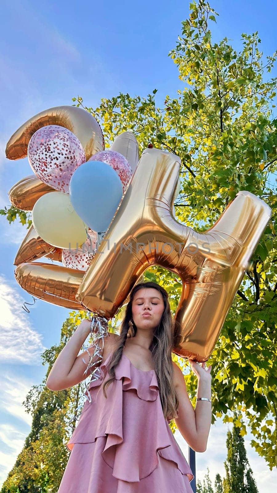Smiling caucasian young girl model holding bunch of golden balloons smiling at camera. Blonde girl is wearing silk dress against blue sky. Birthday and celebration concept. Vertical
