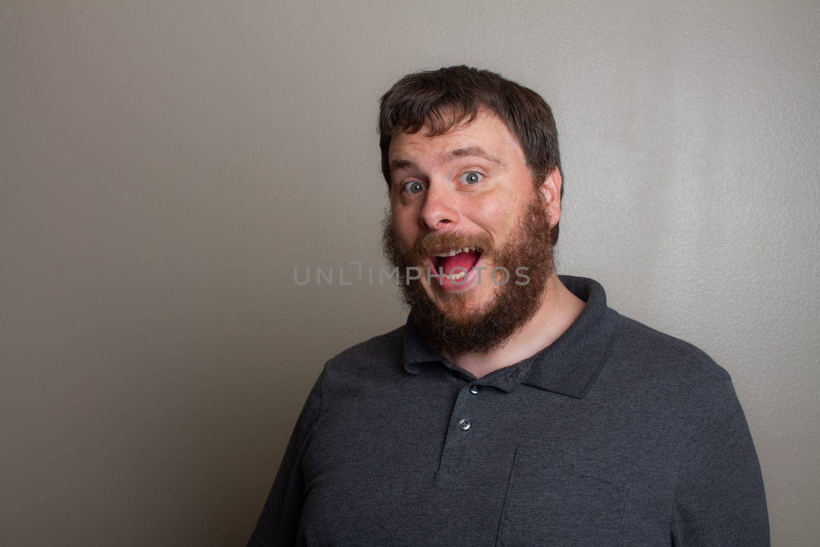A joyful bearded man with an enthusiastic expression, wearing a grey shirt against a plain backdrop. Captures a moment of happiness and surprise, showcasing an exuberant personality and cheerful mood.