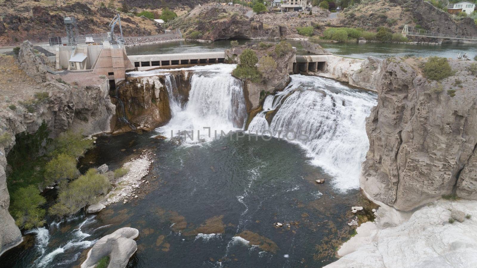 A stunning waterfall cascades over rugged cliffs next to a power plant, set against a scenic landscape. The powerful flow creates a mesmerizing natural scene from shoshone falls in twin falls idaho