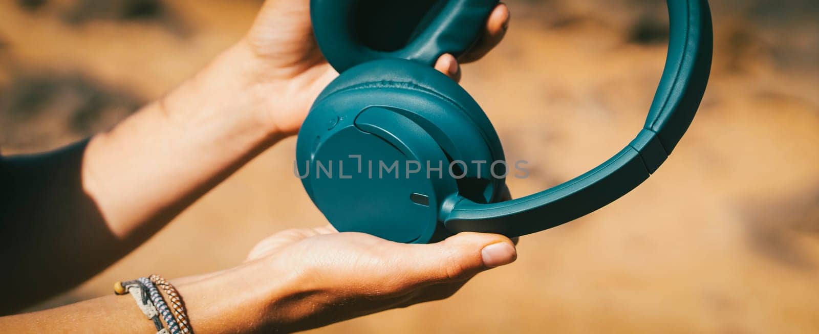 Close-up of female hands with stylish sea-blue wireless headphones against the background of a beautiful ocean with waves.
