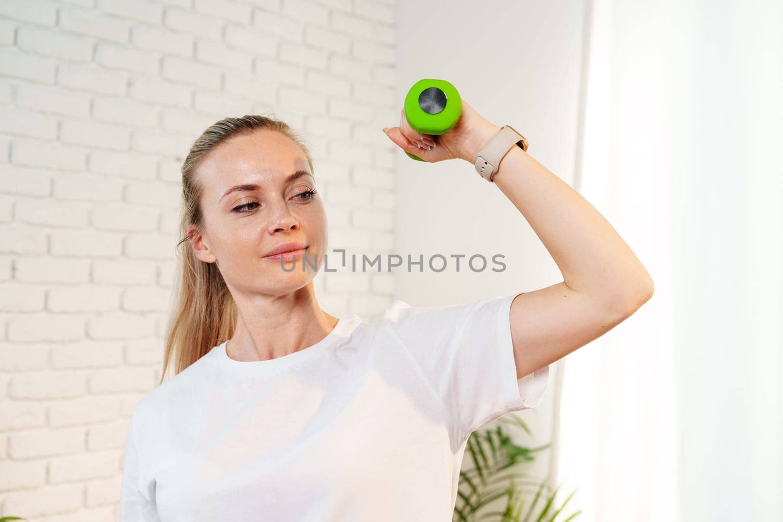 A woman exercises with green dumbbells at home in a bright room filled with light and plants